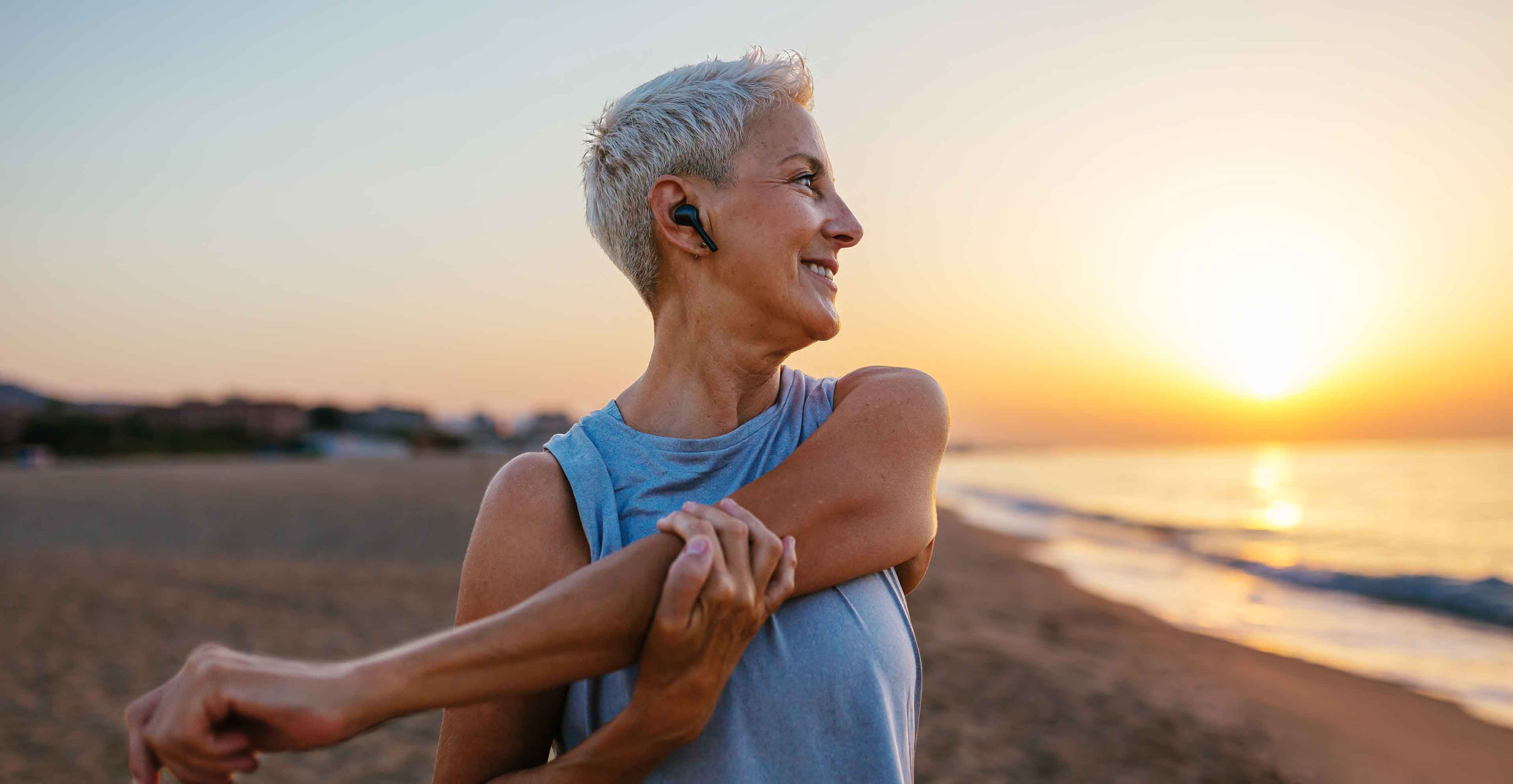 A person stretches their arm on a beach at sunset while wearing wireless earbuds.