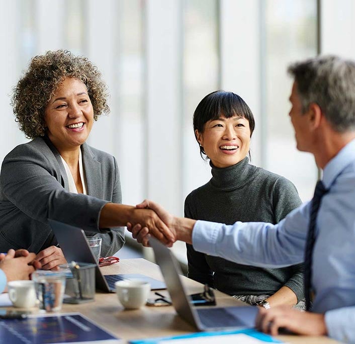People in a business meeting shaking hands across a table with laptops and coffee cups.