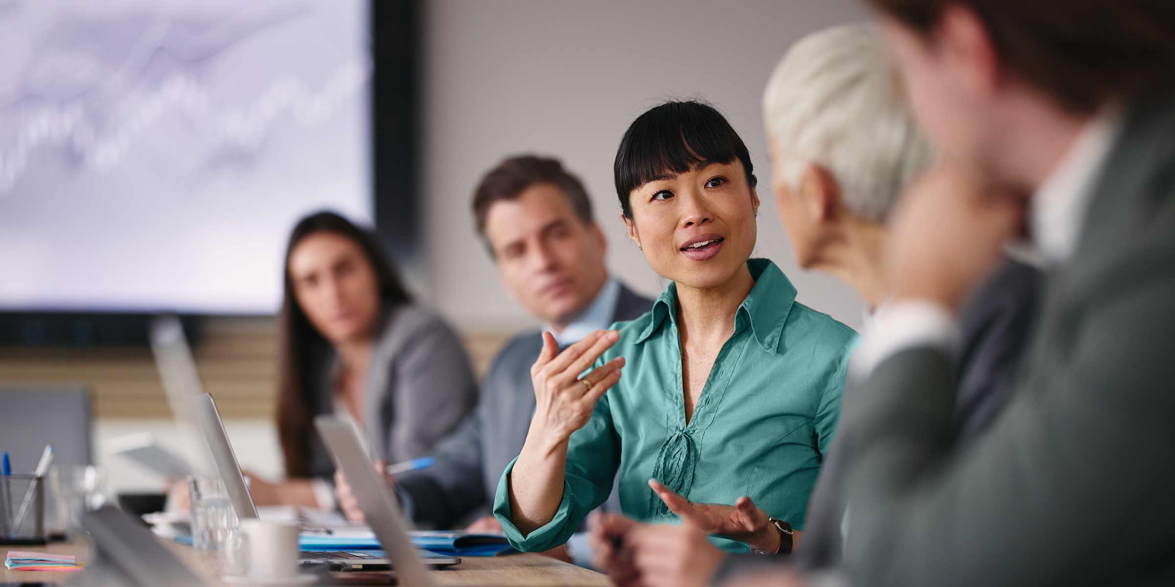 People in a meeting room discussing ideas with laptops and a presentation screen in the background.
