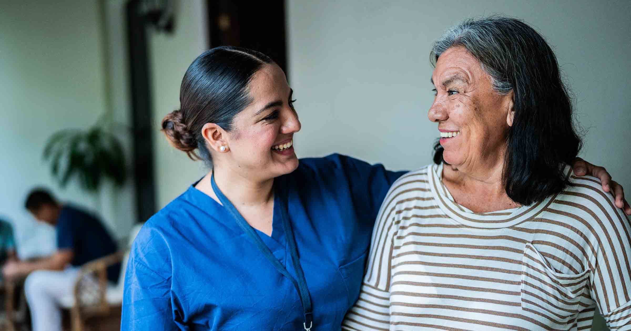 A person in blue scrubs has their arm around another person in a striped shirt indoors.