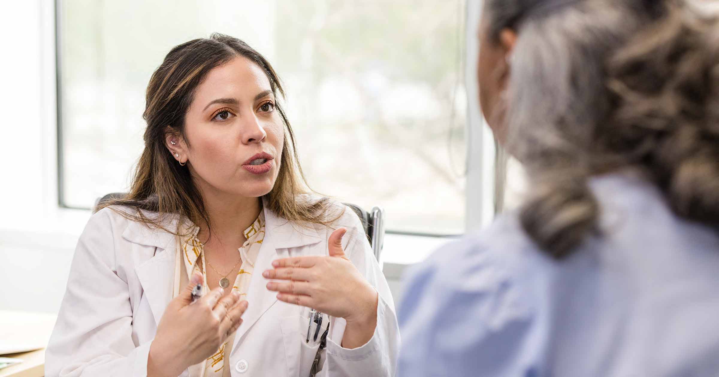 A doctor in a white coat gestures while speaking to a patient in a well-lit office.